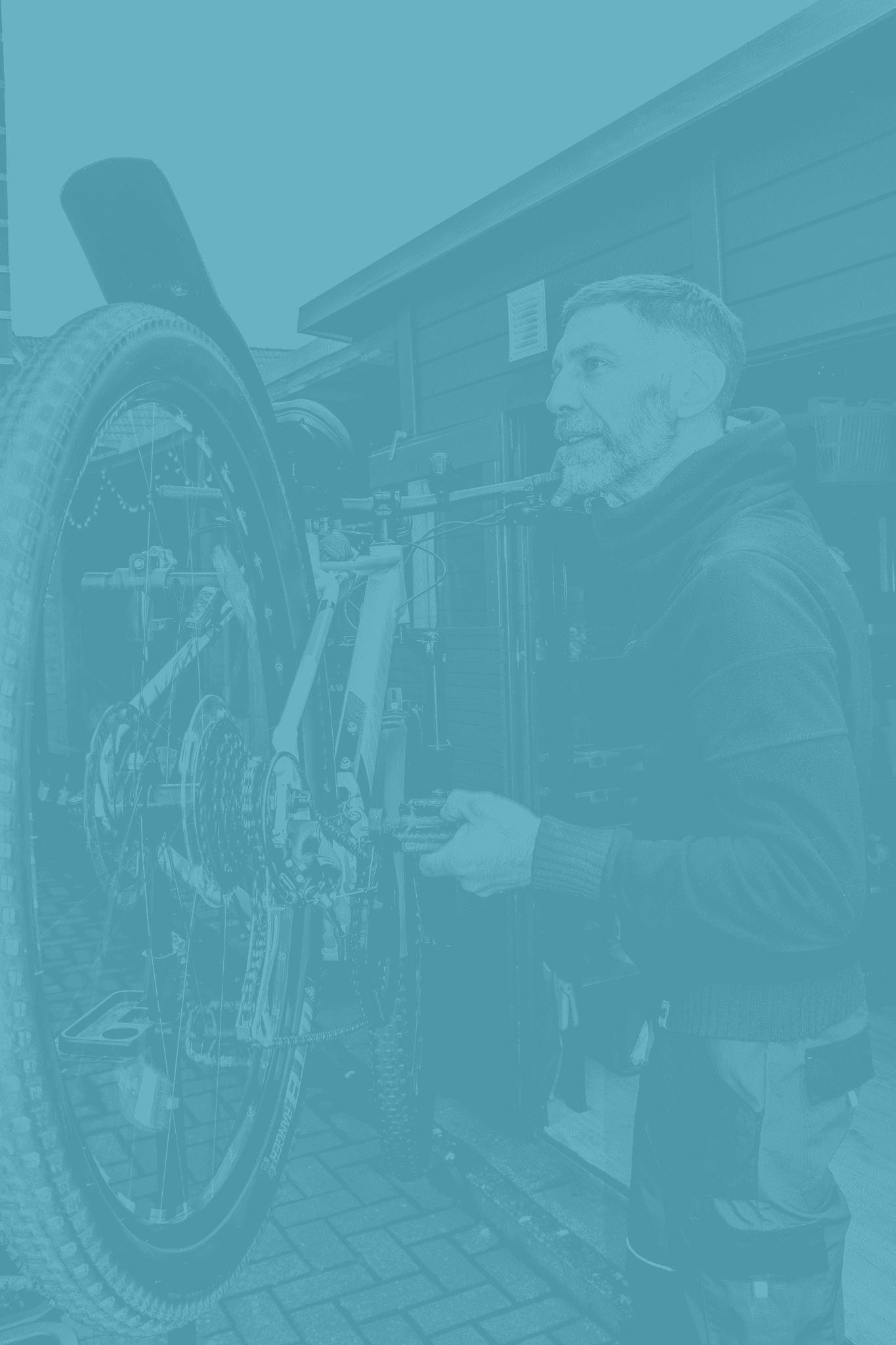 Bärtiger Mann repariert ein senkrecht montiertes Fahrrad an einem Reparaturständer vor einer Werkstatt.