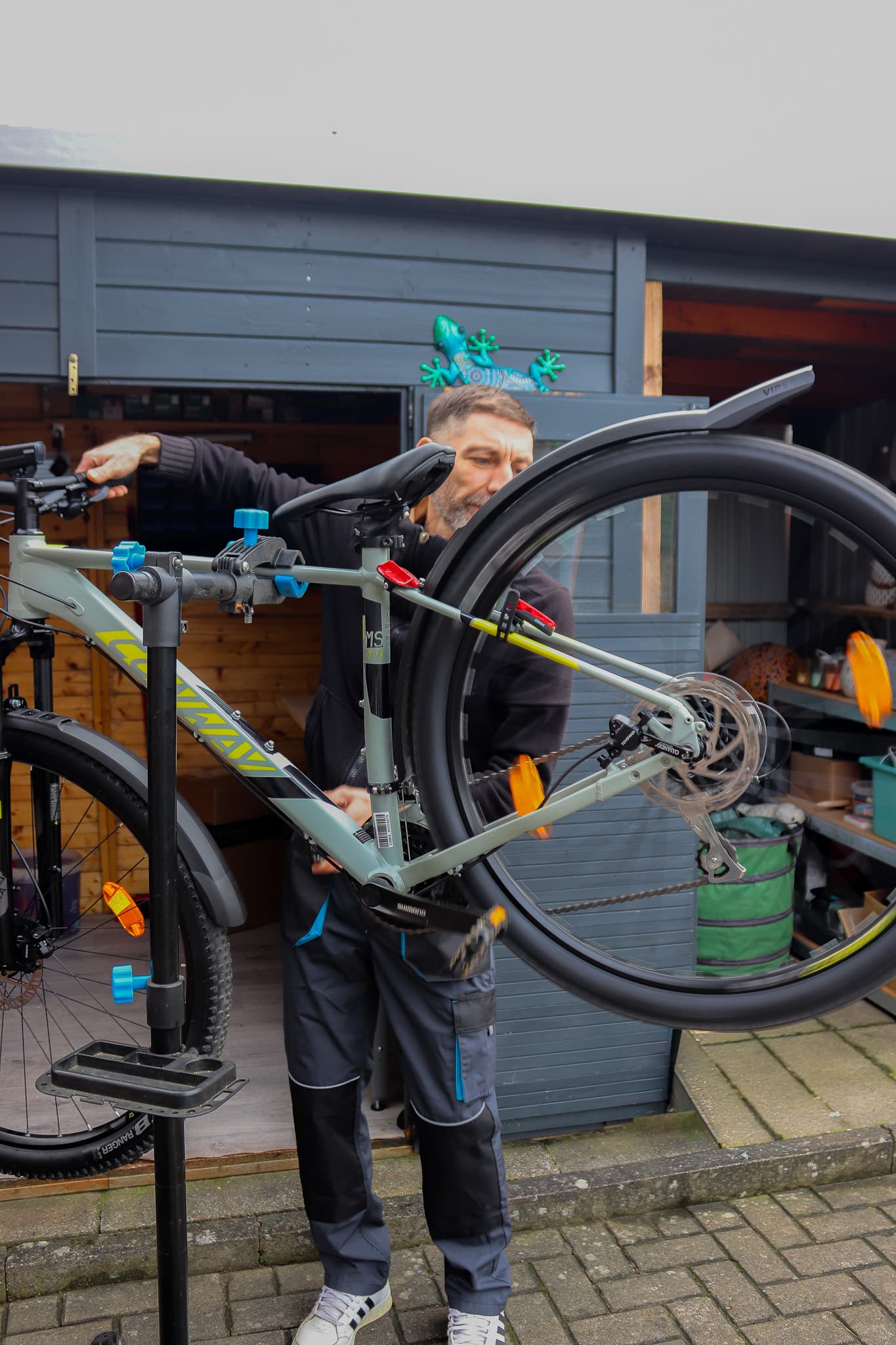 Ein Mann in Arbeitskleidung repariert ein graues Mountainbike an einem Montageständer vor einem blauen Holzschuppen.