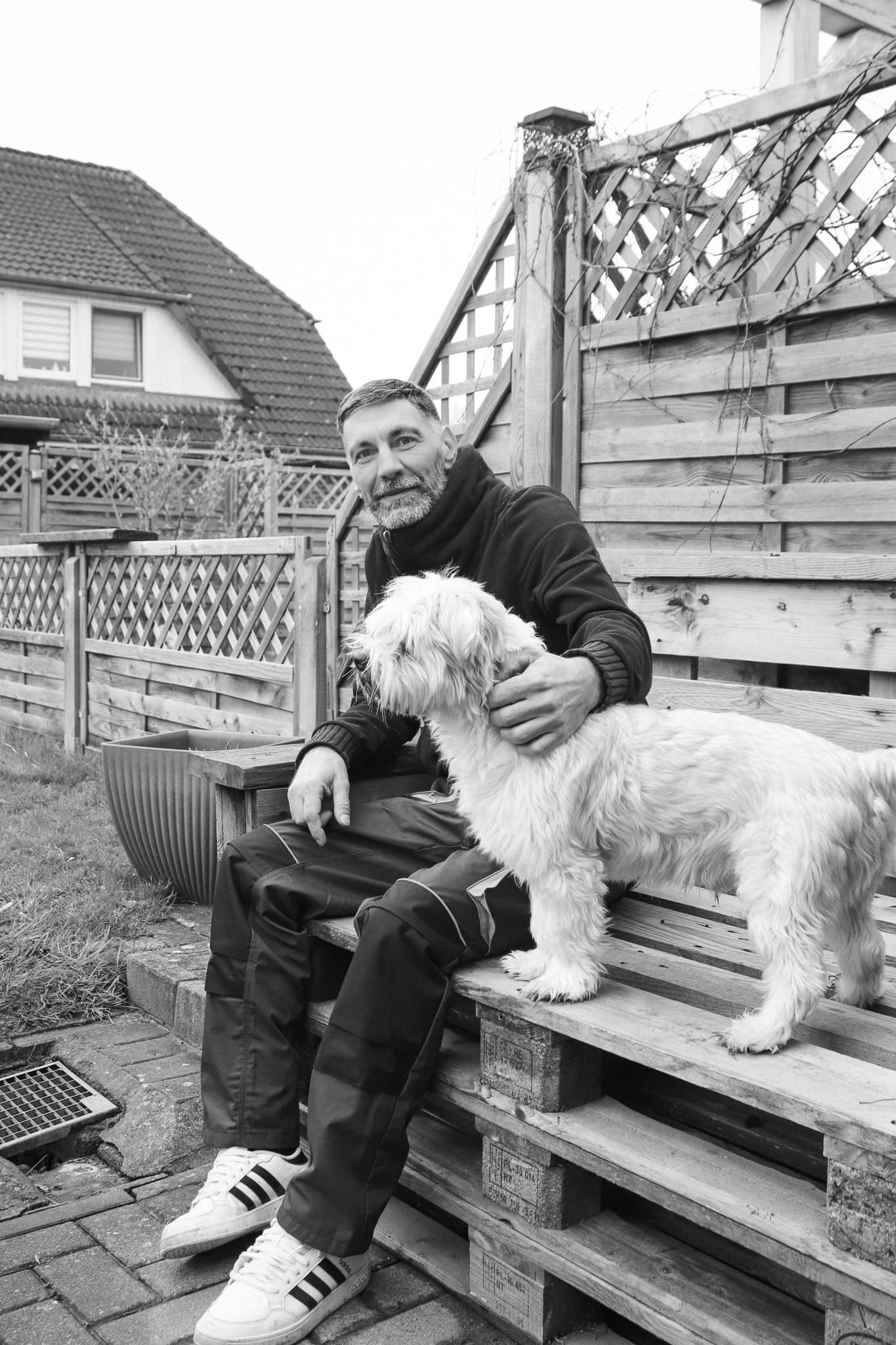 Ein bärtiger Mann sitzt auf einer Palettenbank im Garten und streichelt einen kleinen weißen Hund.