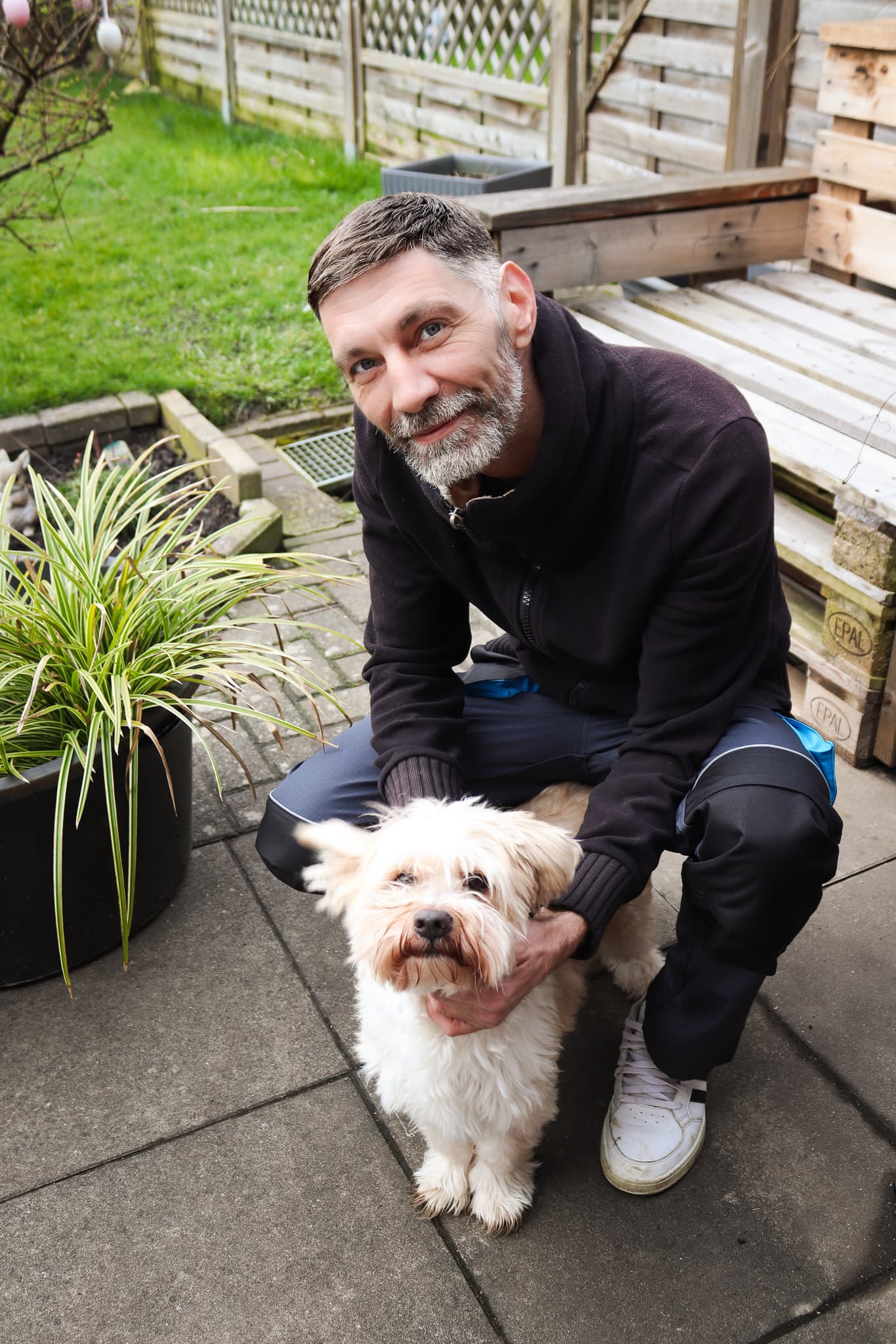 Ein lächelnder Mann mit Bart hockt auf einer Terrasse und streichelt einen kleinen weißen Hund.