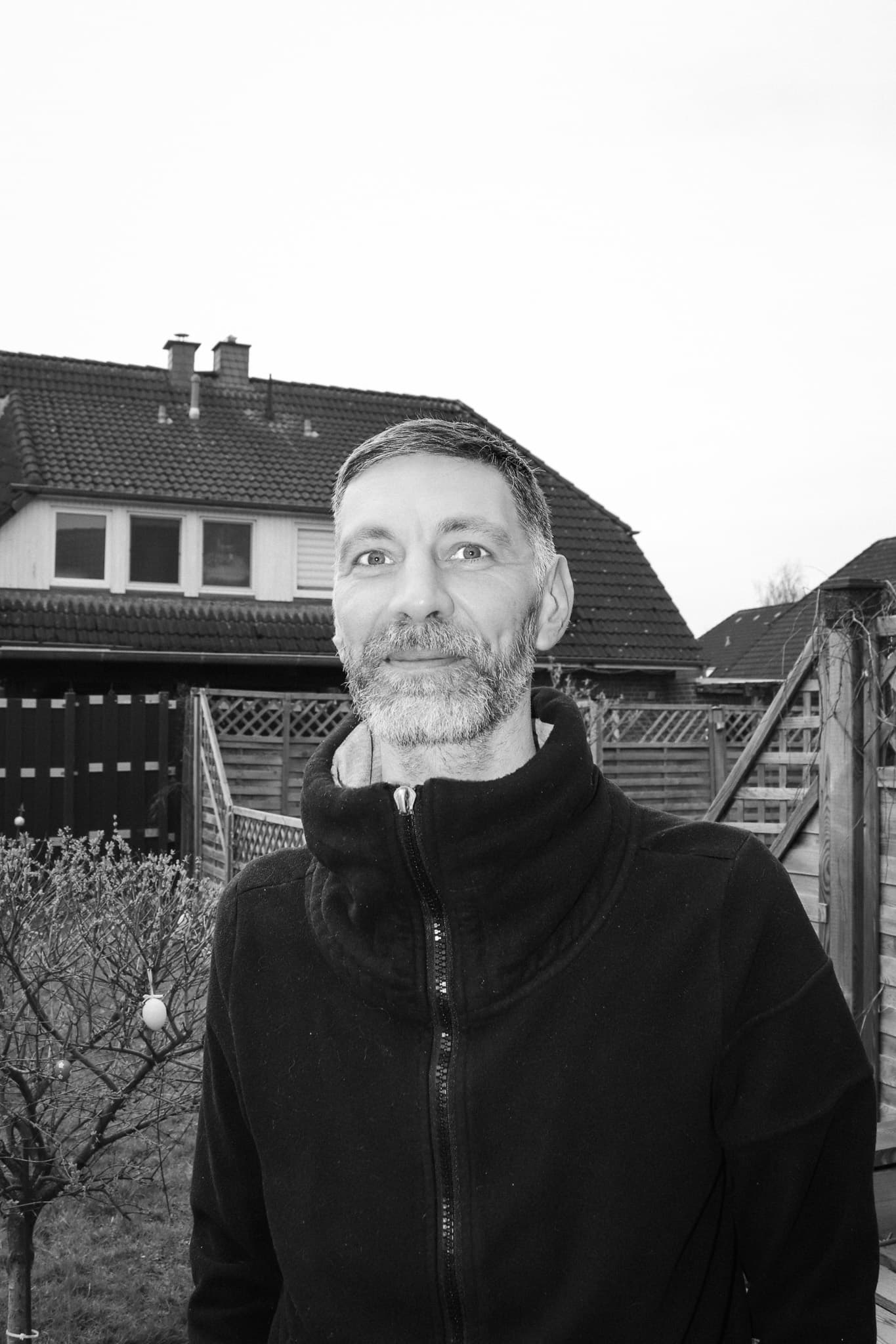 Schwarz-Weiß-Porträt eines lächelnden, bärtigen Mannes in einer dunklen Fleecejacke vor einem Wohnhaus.
