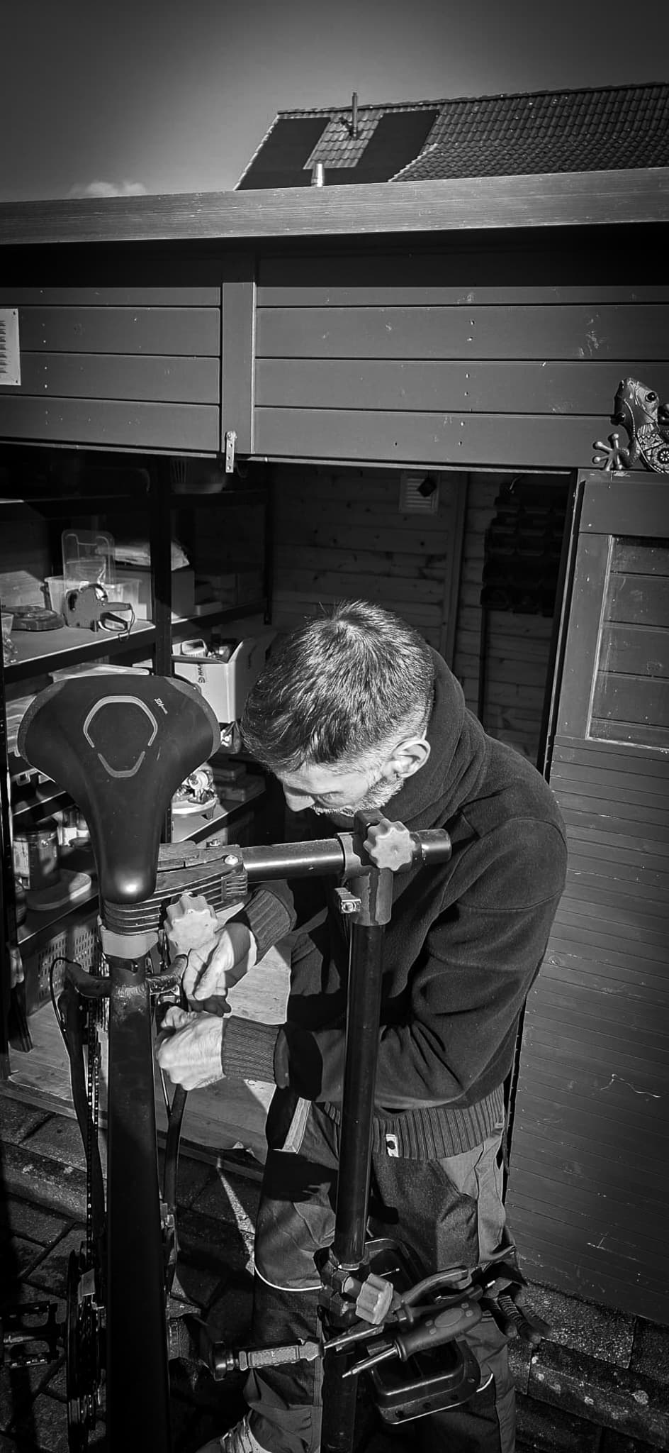 Ein Mann repariert konzentriert ein Fahrrad an einem Montageständer vor einem dunklen Holzschuppen.
