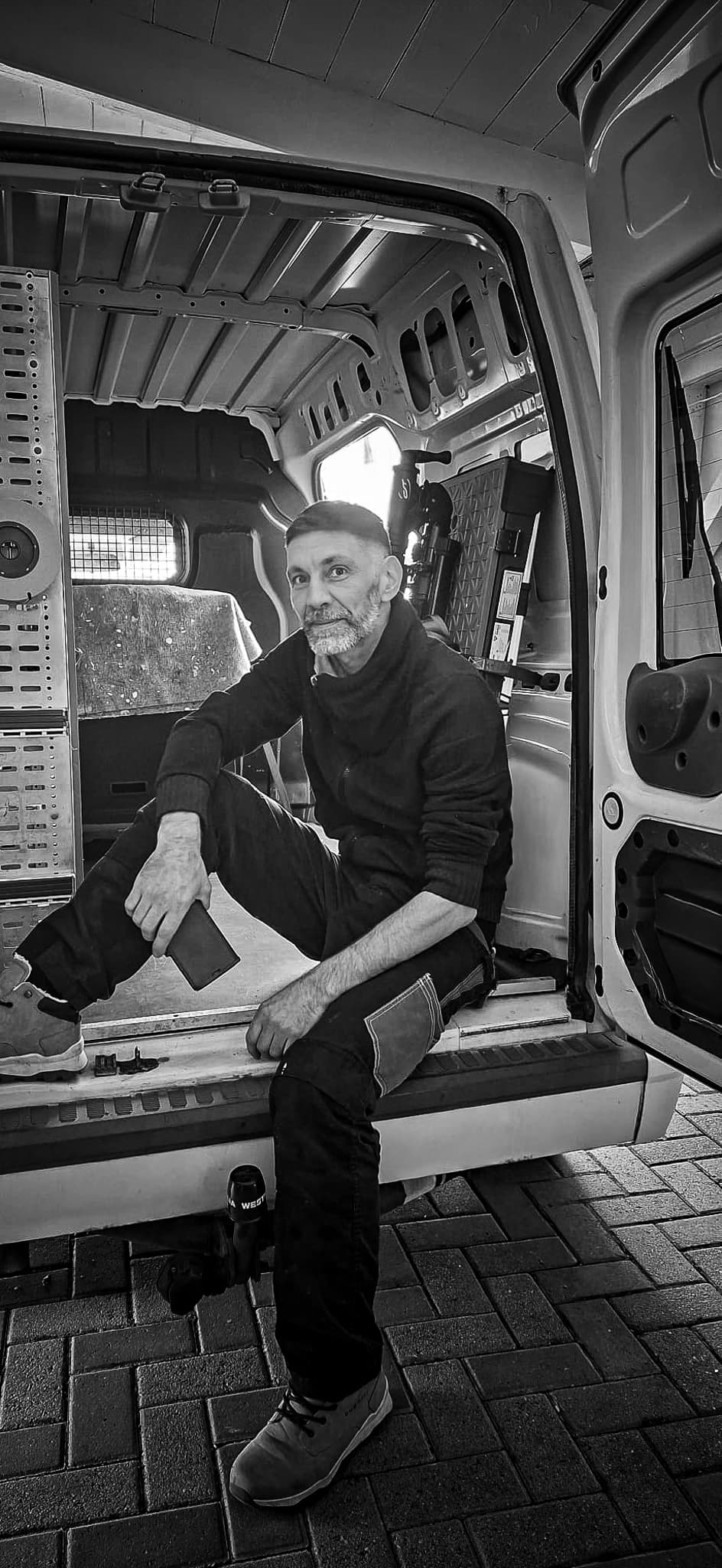 Schwarz-weißes Porträt eines bärtigen Mannes in Arbeitskleidung, der im Heck eines offenen Transporters sitzt.